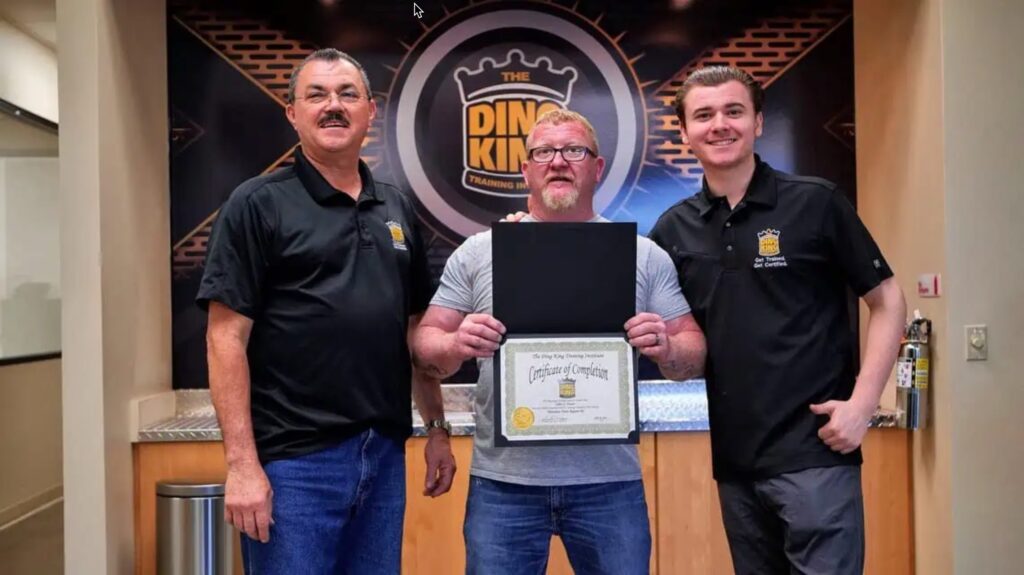 Three men stand indoors in front of a DING KING sign; the man in the middle is holding a certificate of completion for PDR Training in California, and the two men on either side are smiling, wearing matching shirts with the logo.