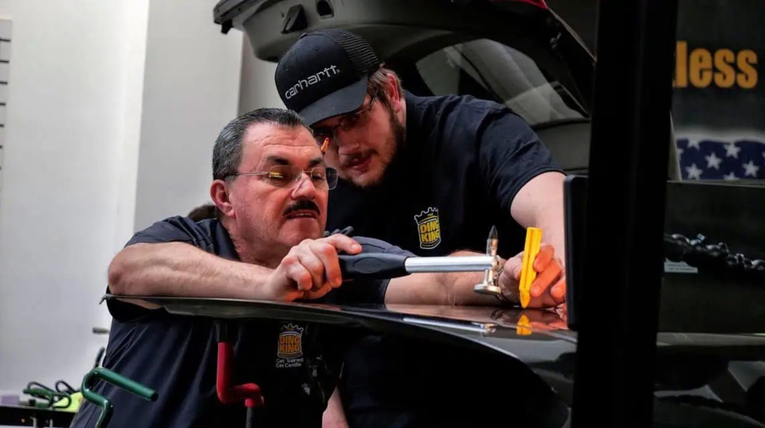 Two men work together to repair a vehicle door dent using specialized tools in a garage. Both wear black shirts with logos and one wears a black cap. They appear focused and engaged in the task, showcasing their skills honed through PDR Training in California.