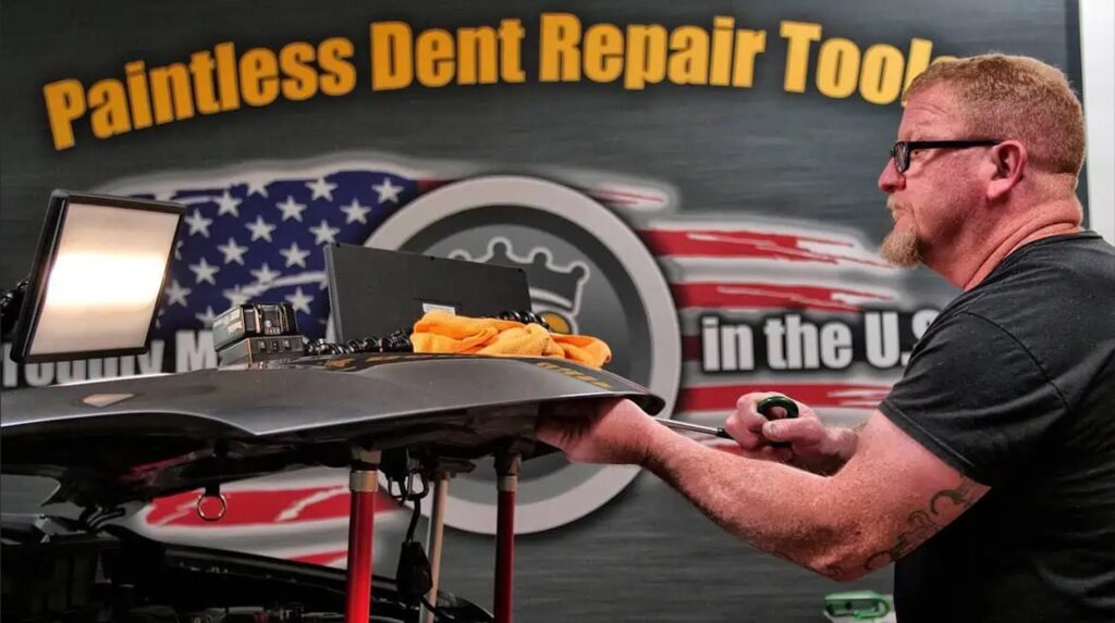 A man works on a car using paintless dent repair tools, with a sign in the background that reads "Paintless Dent Repair Tools" and an American flag design, honing his skills from recent PDR Training in California.