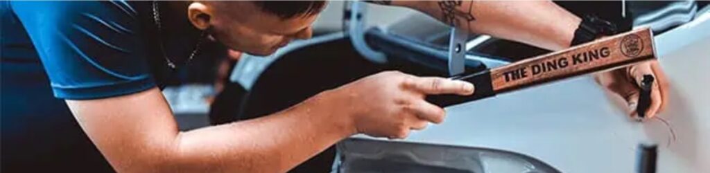 A person in a blue shirt, skilled from PDR Training in California, uses "The Ding King" tool to repair a minor dent on a vehicle's white body panel.