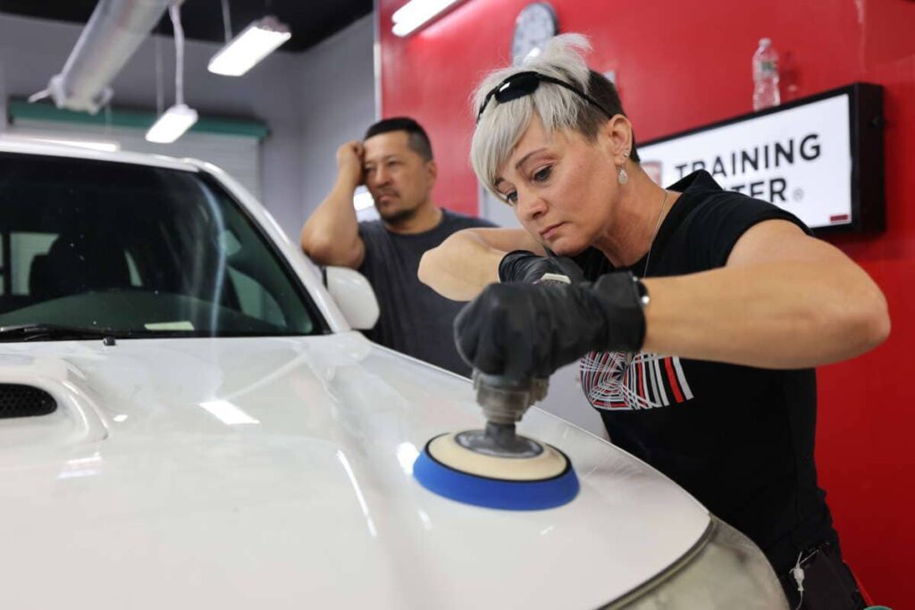 A person with short hair and gloves is using a polishing tool on a white vehicle's hood, likely part of PDR Training in California, while another individual stands in the background watching.