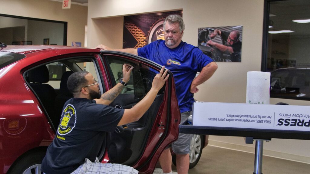 A man kneels beside a red car, applying a window film, while another man stands nearby observing in an indoor garage setting. Both wear branded work shirts from a PDR Training in California session.