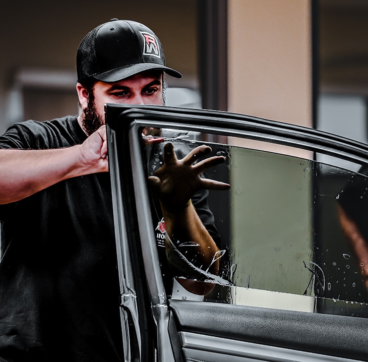 A person wearing a black cap and shirt is applying a film to a partially rolled-up car window, demonstrating techniques from PDR Training in California.