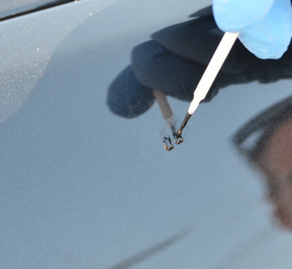 A person wearing a blue glove is meticulously applying touch-up paint to a small scratch on a gray car's surface using a small brush, demonstrating skills often emphasized in PDR training in California.