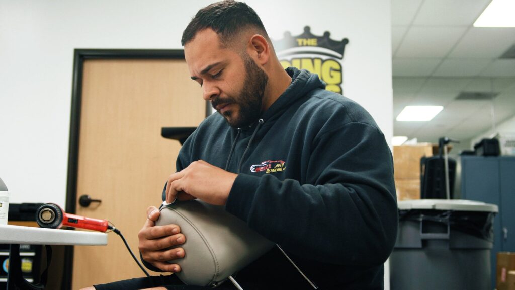 A man in a black hoodie works intently with a tool on a light-colored item in an indoor workspace, focusing on the task at hand. Various equipment and a door can be seen in the background, indicating he might be engaged in PDR Training in California.