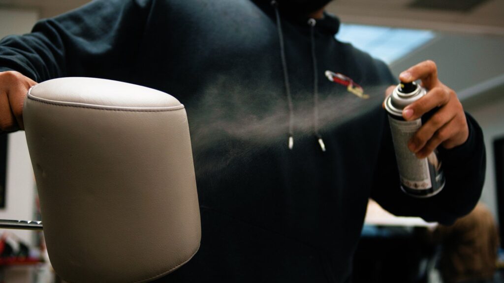 A person in a hoodie is spraying a can of mist onto a padded chair armrest, likely a technique learned from PDR Training in California.