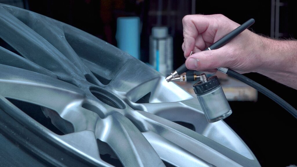 A person is using an airbrush tool to paint or repair a car wheel rim, showcasing skills often learned in PDR Training in California. Various bottles and tools are visible in the background.