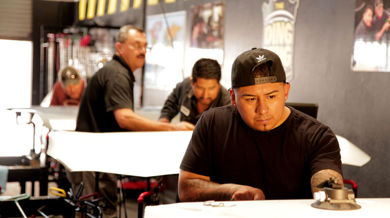 Four men work on repairing vehicle surfaces in an automotive shop. The foreground shows a man intently focused on his task, likely applying techniques learned from PDR Training Courses. Tools and posters are visible in the background.