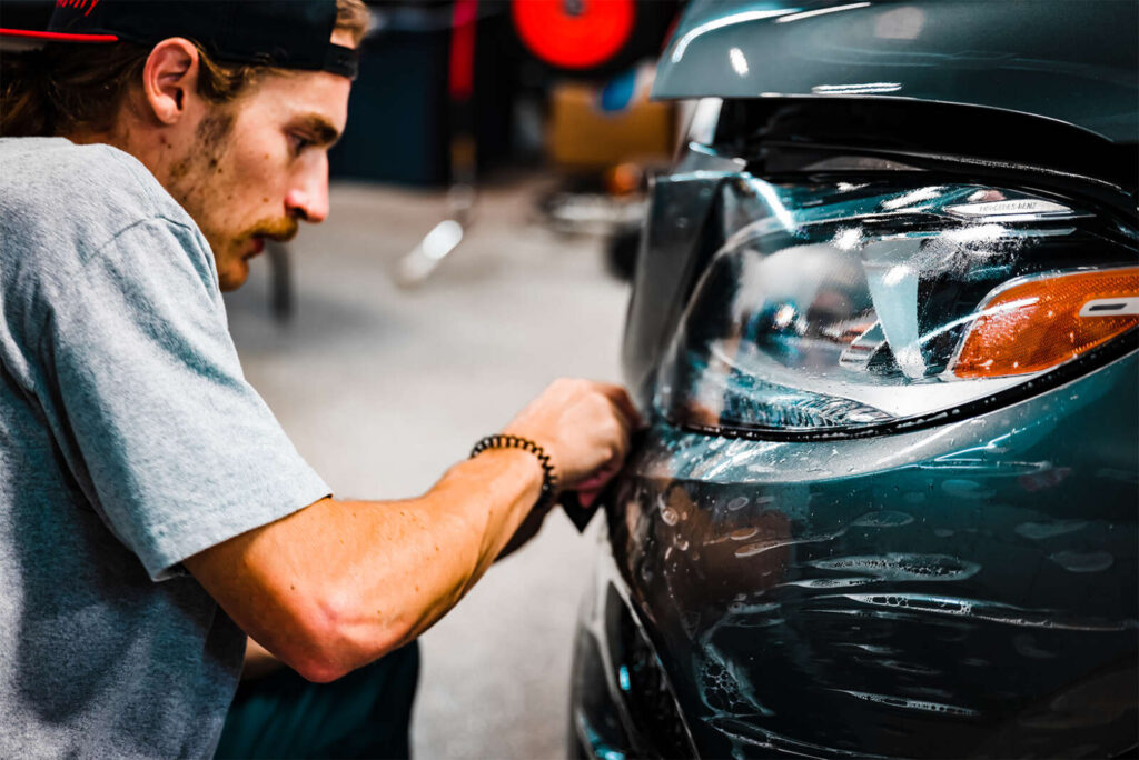 A man in a gray shirt and black cap, evidencing skills from PDR Training in California, repairs the front bumper of a dark-colored car in a garage.