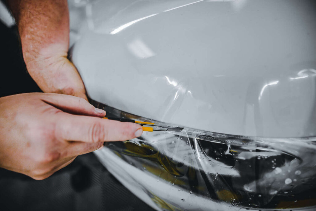 Close-up of hands applying a protective film on a car headlight, using a small plastic tool to smooth out the film—skills that are essential in PDR Training in California.