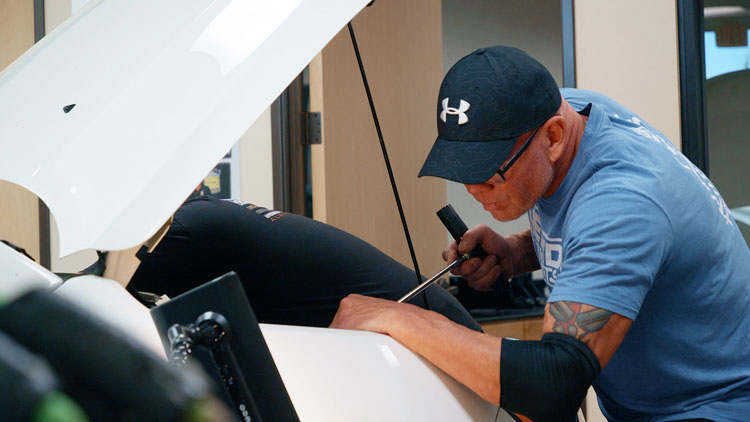 A man in a blue shirt and black cap works on a car with its hood open, using a tool, possibly practicing skills learned from PDR training courses.